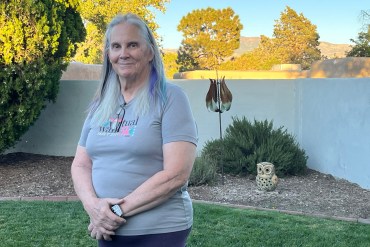 Anne Withrow stands outside near a small garden. A few streaks of her long, gray hair have been dyed purple and blue. She holds her hands together in front of her and looks towards the camera with a slight smile.