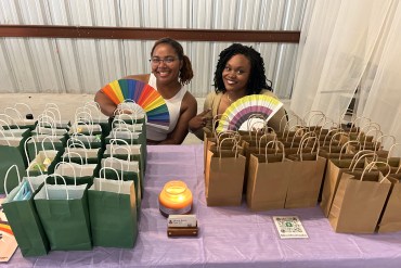 Two college students, Limya Harvey (left) and Cydney Mumford (right), sit at a table covered with small, paper grab-bags. The women smile towards the camera, each holding a paper fan with pride-flag colors.
