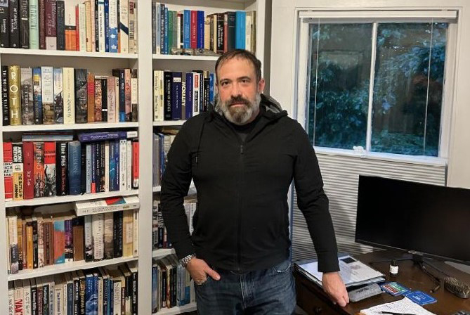 A photo of a man standing for a photo indoors by bookshelves.