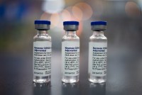 Three vials of the Novavax vaccine are in the center of the image against a bokeh background.