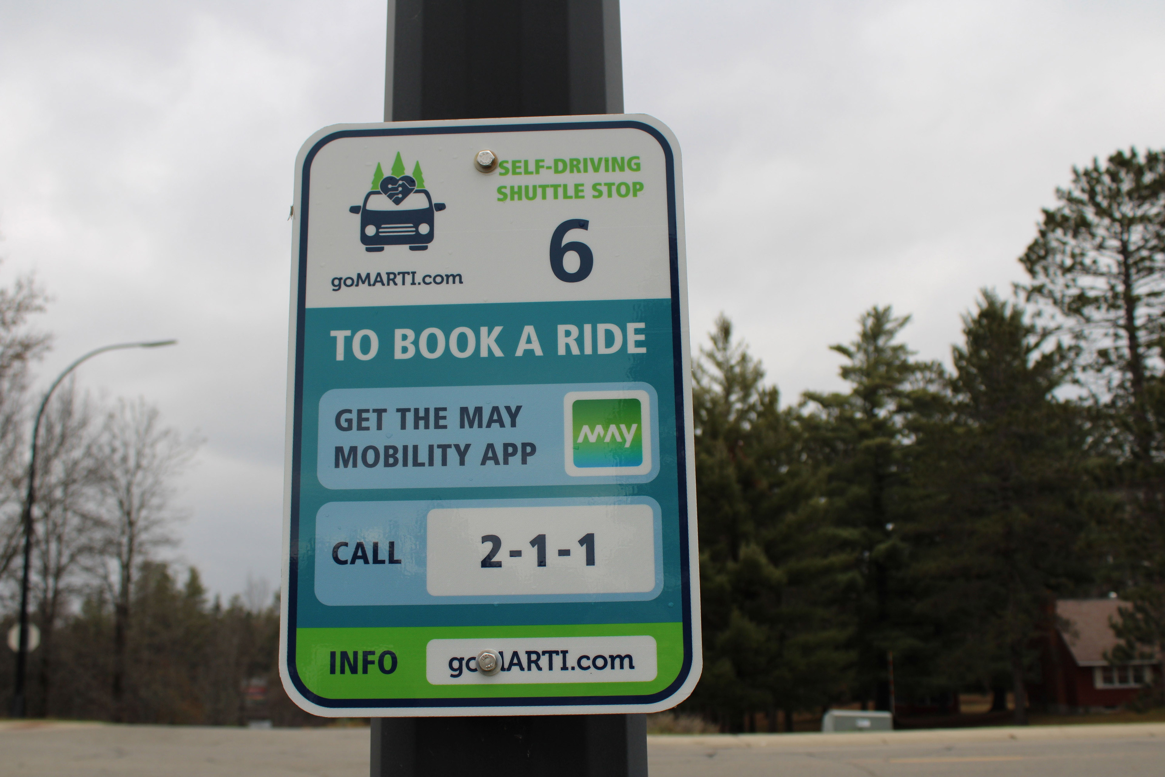 A metal sign is attached to a pole. It has information about how to book a ride for the goMARTI vehicle. It reads, "Self driving shuttle stop 6 / To book a ride get the may mobility app / call 2-1-1 / info: goMARTI.com"