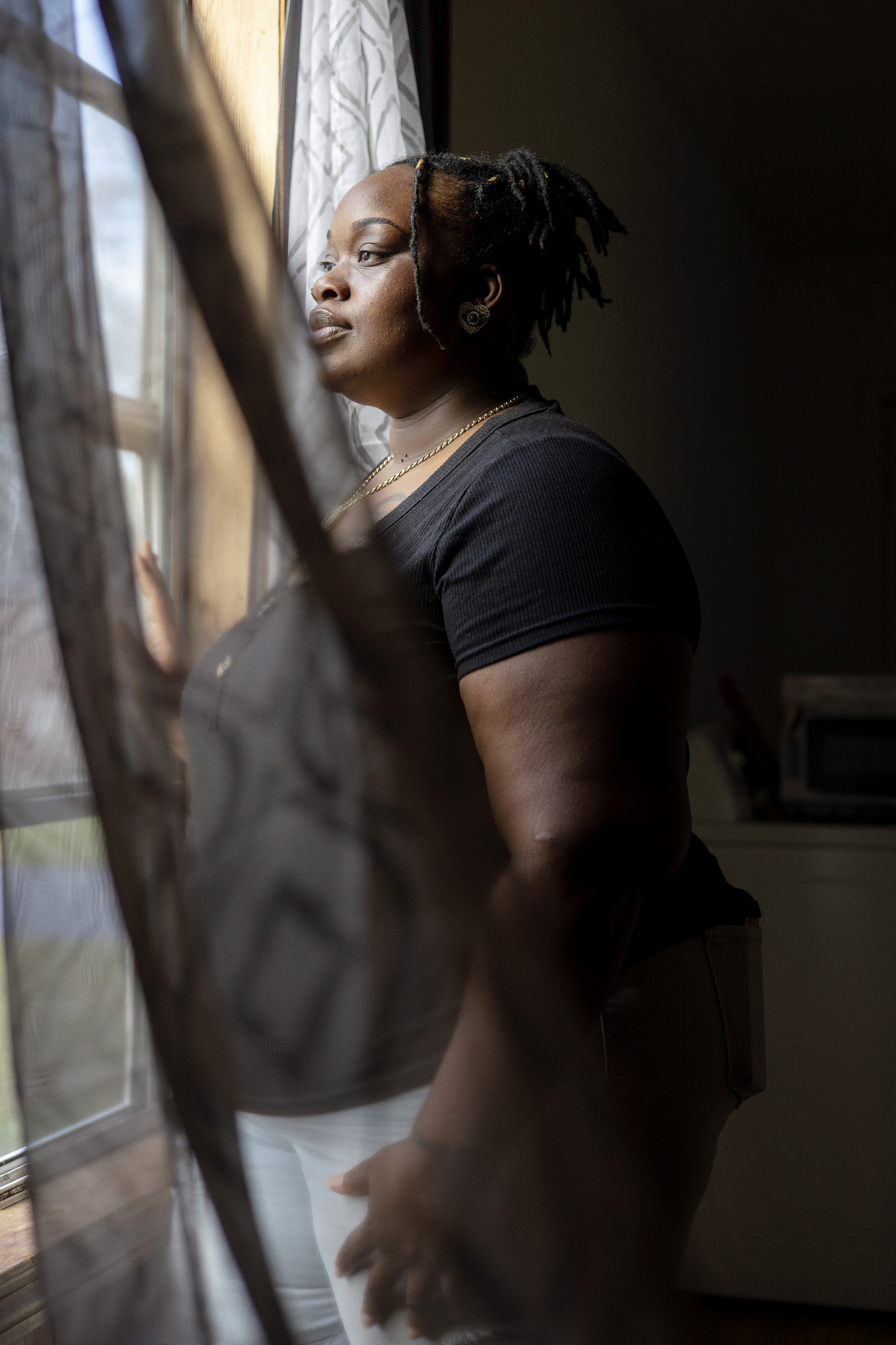 O'laysha Davis looks out a window in her home. A sheer curtain blows in the wind in front of her figure.