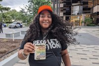 Shameka Parrish-Wright stands in the center of the photo, facing the camera with a smile. She is visible from the waist up, and holds a lanyard tag that in front of her that reads, "House is healthcare / public safety / education /infrastructure / family values / stability / welcome / public health / a human right / first."
